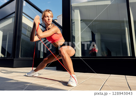 Woman performing resistance band exercise outdoors near a modern glass building Woman performing resistance band exercise outdoors near a modern glass building 128943043