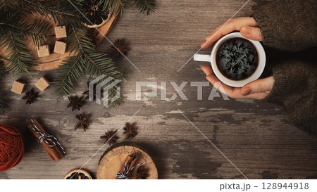 Christmas flat lay. Female hands in a cozy green sweaters sleeves with coffee cup and frosty patterns in it. Spruce twigs, anise, brown sugar, a ball of wool on a wooden table. 128944918