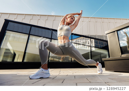 Slender blonde sportswoman doing exercises against the blue summer sky and gray modern building background. Young female athlete enjoys morning workouts. Regular trainings in urban environment. Slender blonde sportswoman doing exercises against the blue summer sky and gray modern building background. Young female athlete enjoys morning workouts. Regular trainings in urban environment. 128945227