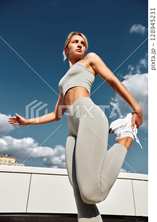 Young female athlete doing outside workout on a sunny day. Slender blonde girl in gray sportswear makes jumps, does functional training in the street. Active lifestyle in urban environment. Young female athlete doing outside workout on a sunny day. Slender blonde girl in gray sportswear makes jumps, does functional training in the street. Active lifestyle in urban environment. 128945231