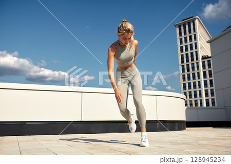 Young female athlete doing outside workout on a sunny day. Slender blonde girl in gray sportswear makes jumps, does functional training in the street. Active lifestyle in urban environment. 128945234