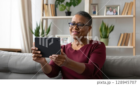 Cheerful senior woman video chatting on digital tablet in cozy home setting with bookshelves. Generative AI. 128945707