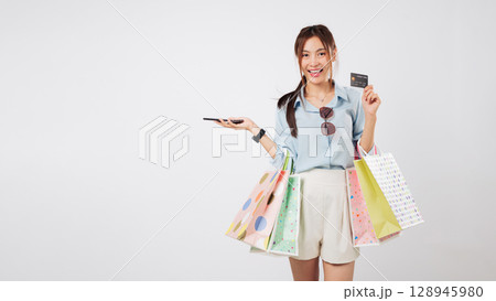 Happy asian woman with bag shopping online, holding phone and credit card, feeling excited, showing a card with shopping bags after successful mobile payment, headshot isolated studio white background 128945980