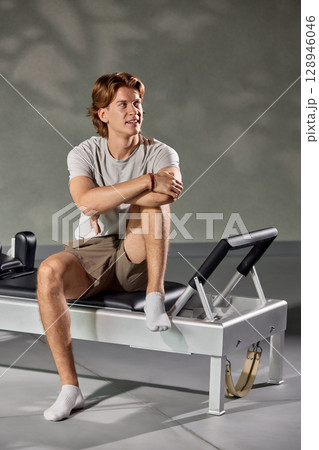 Man seated on a Pilates reformer machine in a fitness studio Man seated on a Pilates reformer machine in a fitness studio 128946046