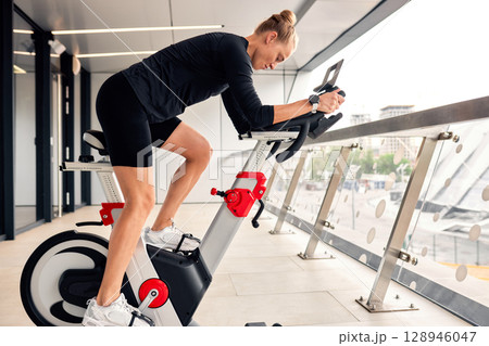 Woman exercising on a stationary bike indoors with a city view. 128946047