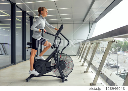 Woman exercising on a modern air bike in a sleek gym interior Woman exercising on a modern air bike in a sleek gym interior 128946048