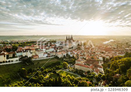 Golden sunlight bathes Prague Castle and the surrounding city as day breaks. The colorful rooftops and lush greenery create a captivating start to the day in this historic location. 128946309