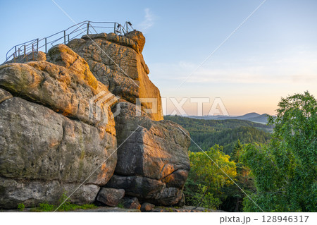 Golden sunlight bathes the sandstone cliffs of Popov Cliffs in Czechia during a tranquil evening. Lush greenery surrounds the striking rock formations in this serene landscape. 128946317