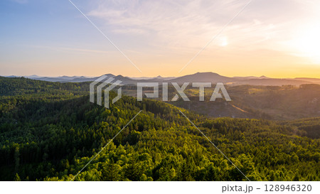 A breathtaking sunset illuminates the Lusatian Mountains from the vantage point of Popov Cliffs. The landscape features lush green forests, rocky formations, and distant peaks. 128946320