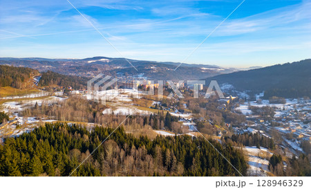 Aerial view captures the snowy landscape of Tanvald in the Jizera Mountains, showcasing the serene beauty of winter with rolling hills, forests, and distant buildings. Aerial view captures the snowy landscape of Tanvald in the Jizera Mountains, showcasing the serene beauty of winter with rolling hills, forests, and distant buildings. 128946329