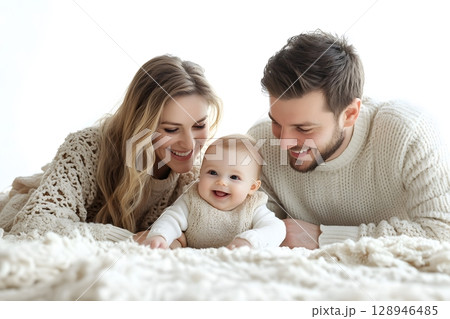 Joyful mother, father, and their sweet baby lie on a soft, light-colored blanket, all smiling in a loving family moment. 128946485