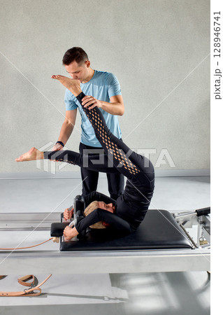 Instructor assisting a pilates exercise on a reformer machine in a gym setting 128946741