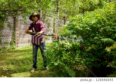 Man skillfully trimming bushes with a hedge trimmer on a sunny day. 128946843