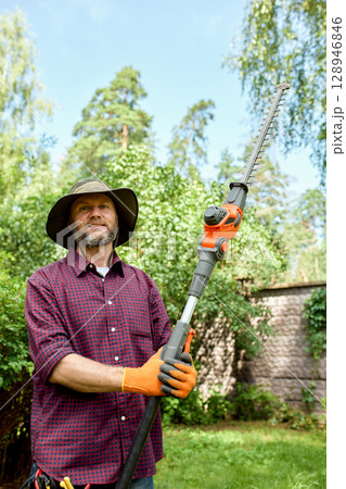 Man trimming garden hedge with electric trimmer on a sunny day outdoors 128946846