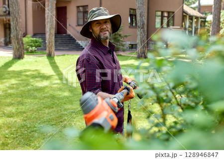 Man tending garden with electric trimmer in backyard 128946847