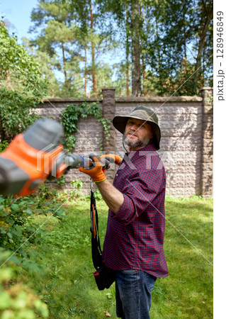 Man trimming hedges in a garden with electric tool on a sunny day Man trimming hedges in a garden with electric tool on a sunny day 128946849