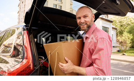 Man Loading Groceries From a Paper Bag Into a Car Trunk Outdoors Man Loading Groceries From a Paper Bag Into a Car Trunk Outdoors 128946857