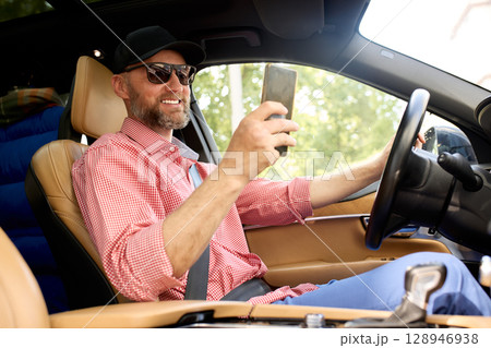 Man driving a car while holding a smartphone and smiling in sunlight Man driving a car while holding a smartphone and smiling in sunlight 128946938