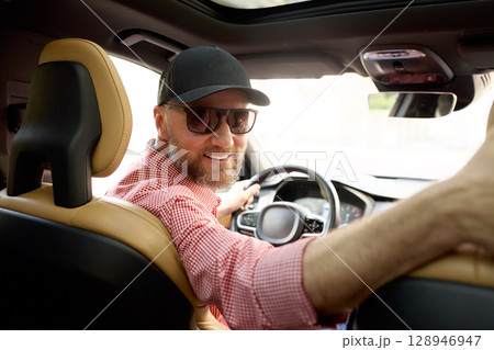 Cheerful man driving a modern car while glancing toward the backseat Cheerful man driving a modern car while glancing toward the backseat 128946947