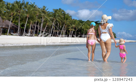 Rear view of mom and daughters walking in warm ocean water on sunny tropical beach 128947243