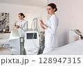 Professional cosmetologist in white uniform with modern aesthetic laser machine for non-invasive skin treatment in beauty clinic or medical spa, advanced cosmetic equipment in clean bright interior 128947315