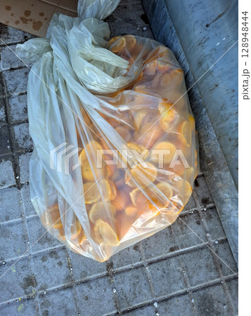 A sizable bag containing numerous orange peels sits on the pavement after the oranges have been juiced. The setting appears to be in a public space, possibly near a market or food area. 128948444
