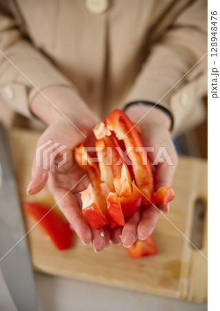 A woman's hands hold pieces of sliced red sweet pepper, top view, a wooden chopping board and a kitchen knife lie in the background 128948476