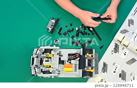 A child's hands assembling a LEGO car model. Various colorful pieces are scattered on a green surface.  128948711
