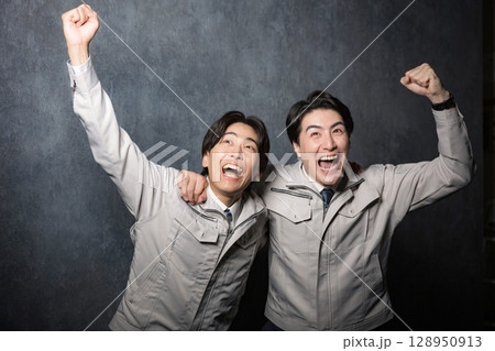 グレーバックで作業着の若い男性のガッツポーズで肩を組んで喜ぶ2人 128950913