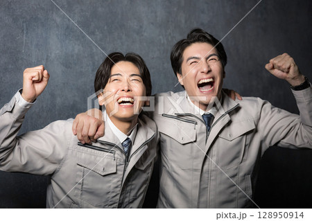 グレーバックで作業着の若い男性のガッツポーズで肩を組んで喜ぶ2人 グレーバックで作業着の若い男性のガッツポーズで肩を組んで喜ぶ2人 128950914