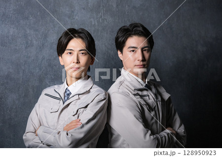 背中合わせで作業着でカメラ目線の真剣な表情がグレーバックの男性2人 背中合わせで作業着でカメラ目線の真剣な表情がグレーバックの男性2人 128950923