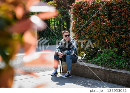Russian male tourist resting on a stone bench near autumn shrubs in a city park, enjoying a sunny day during his travels. Russian male tourist resting on a stone bench near autumn shrubs in a city park, enjoying a sunny day during his travels. 128953081