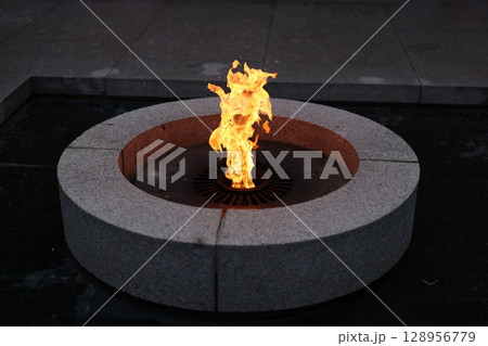 A flame flickers in a circular stone structure, creating a captivating nighttime atmosphere 128956779
