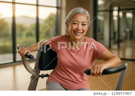 AI generated image of an elderly Asian woman in her 60s exercising in a spacious gym with natural light. She is smiling while using equipment, promoting healthy aging and well-being. 128957467