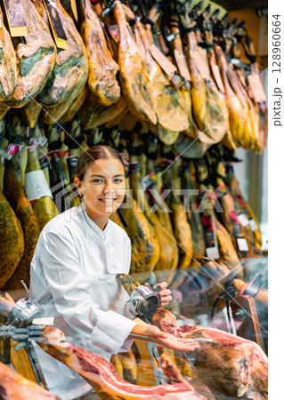 Young girl seller in white jacket selling dry-cured jamon in butcher shop 128960664