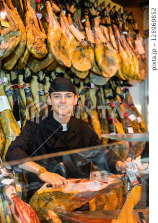 Male seller cutting slices from whole leg of jamon in meat store 128960852