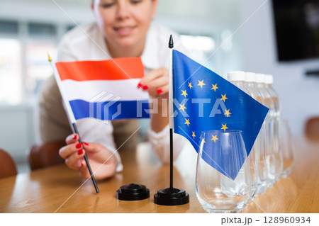 Young woman putting flags of Netherlands and EU on table in office 128960934