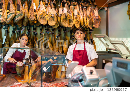 Young man and woman selling spanish jamon at counter Young man and woman selling spanish jamon at counter 128960937
