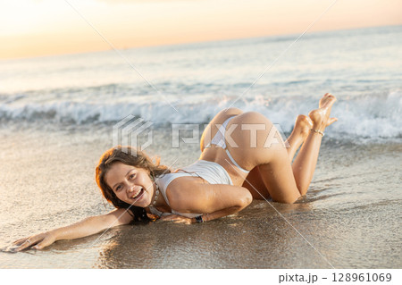 Young woman in thong and top posing while kneeling on beach Young woman in thong and top posing while kneeling on beach 128961069