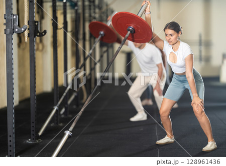 Young woman training with barbell with one arm 128961436