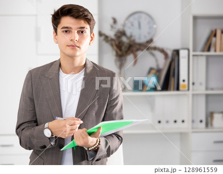 Portrait of man entrepreneur standing in his office, holding paper folder 128961559