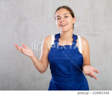 Portrait of young woman builder or mechanic in blue protective overalls on gray background 128961589