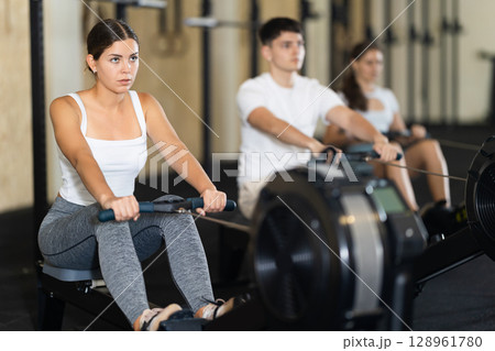 Workout girl cross training exercising cardio using rowing machine with group of people in crossFft club 128961780
