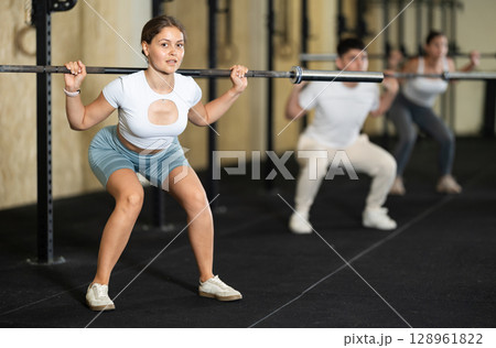 Girl in activewear exercising with barbell during group workout in gym center 128961822