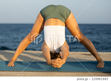 On windy day young slender woman does yoga near sea doing shirshasana 128961885