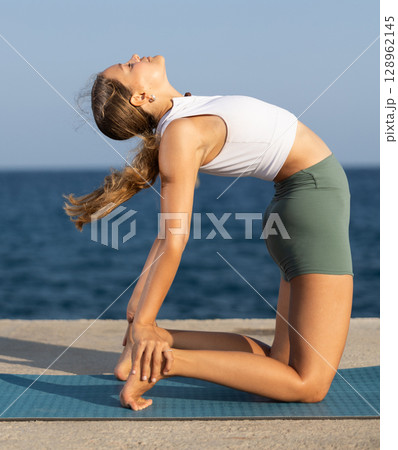 Sportive girl practicing camel pose of yoga on seashore 128962145