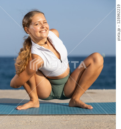 Sportive girl practicing frog pose of yoga on seashore 128962219