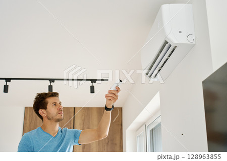 Man using remote control to adjust temperature on wall-mounted air conditioner in modern home interior. Concept of home cooling and indoor climate control during hot summer weather 128963855