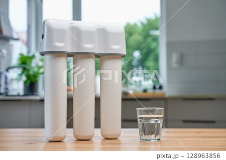 Glass of clean water on wooden kitchen table with water filtration system in background. Purified drinking water for healthy lifestyle 128963856
