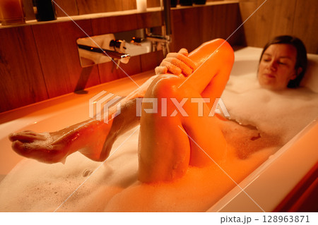 Woman lying in bathtub filled with foam and washing her legs. Female relaxing in bathroom with candles and ambient lighting Concept of wellness, relaxation, and self care 128963871